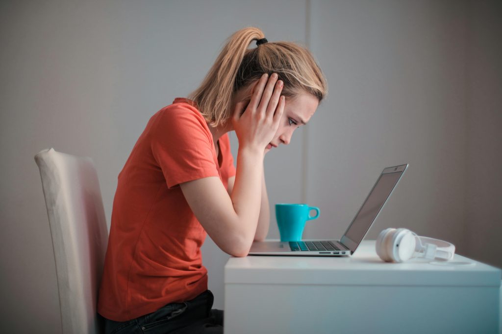 A woman sits indoors at a table, looking concerned while using a laptop with headphones and a coffee mug nearby.